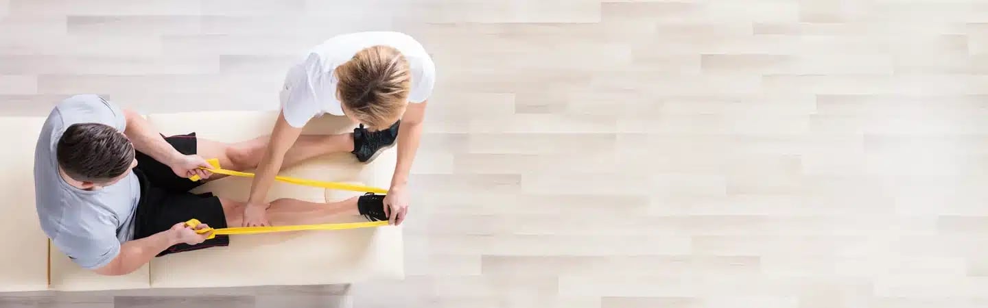 A person helps another stretch their leg with a yellow resistance band on a light wooden floor. Top-down view.