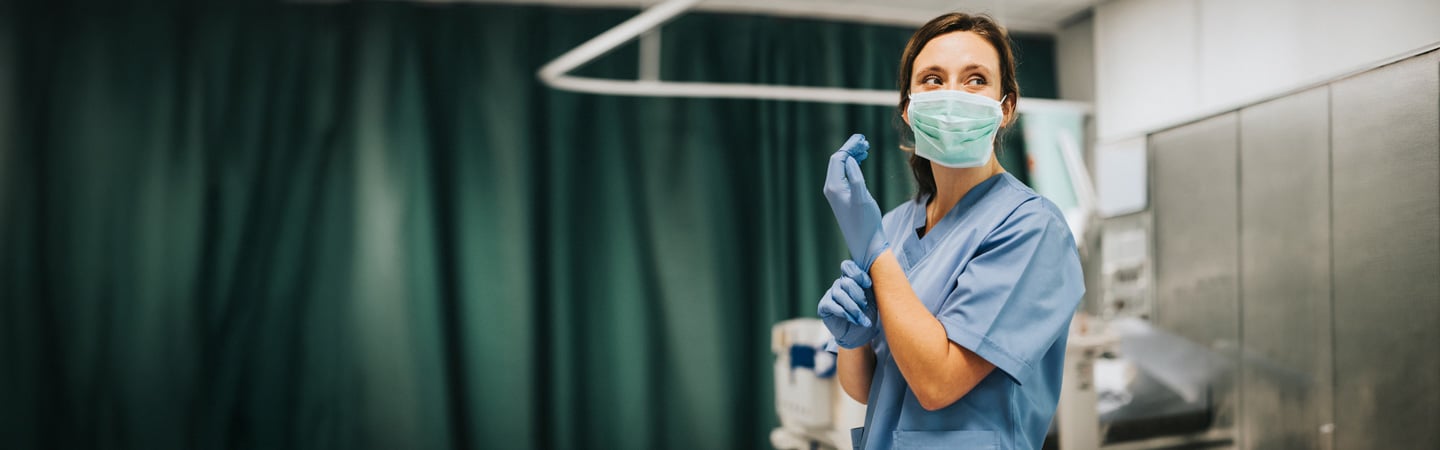 A healthcare worker wearing scrubs, a surgical mask, and gloves stands in a hospital room with green privacy curtains, preparing for a medical procedure.