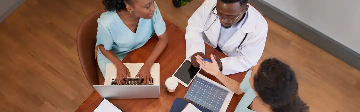 Three medical professionals discussing and reviewing documents around a table with laptops and tablets.