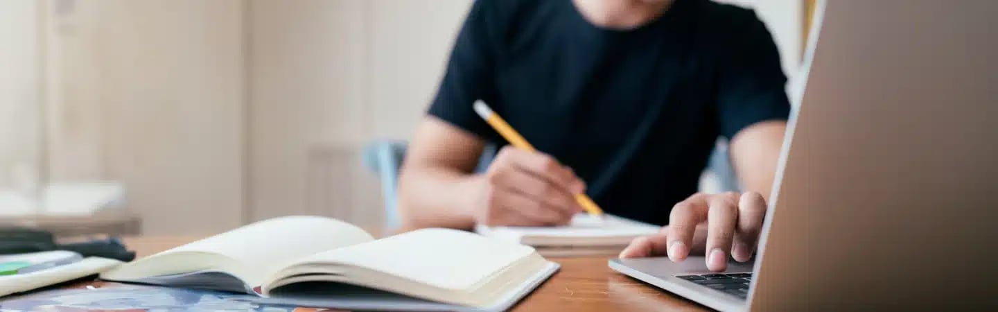 Person writing in a notebook while using a laptop, with books and a pencil on a wooden desk.