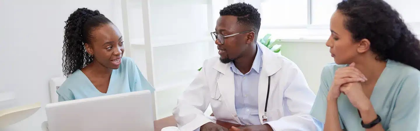 Three medical professionals having a discussion at a table with a laptop in a bright office setting.