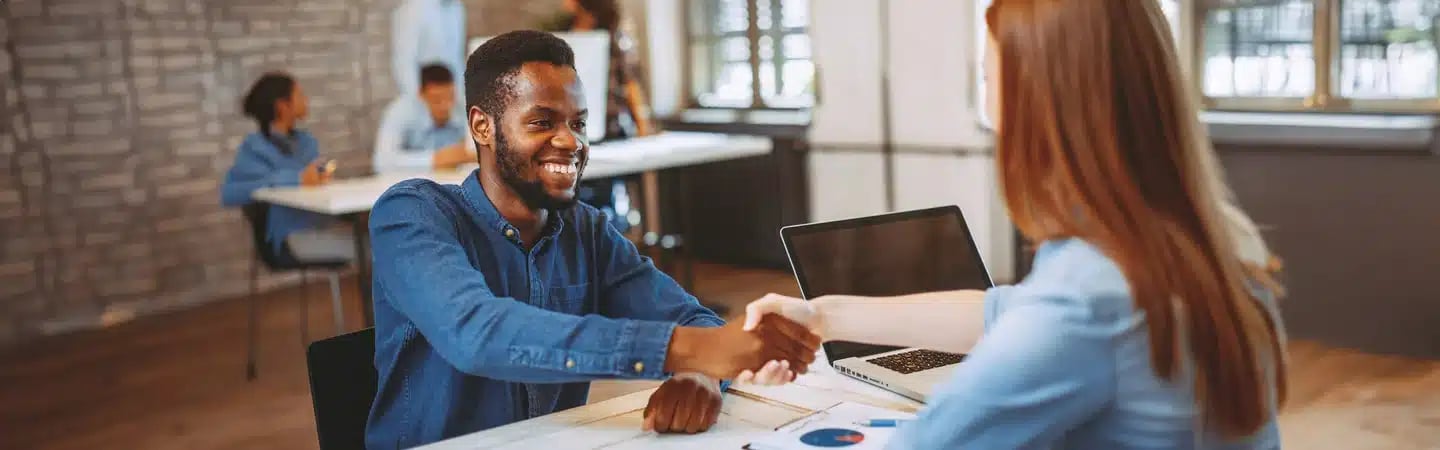 Two people shake hands across a desk in an office setting, smiling. Others work in the background.