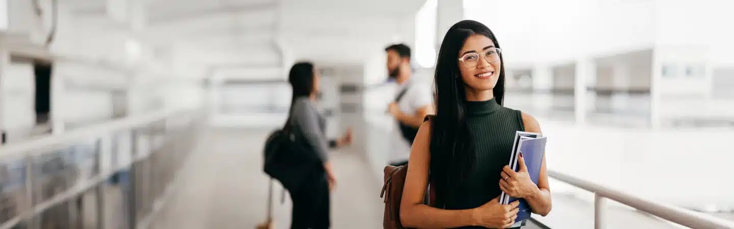 Smiling woman with long hair and glasses holding books, standing in a bright space with people in the background.