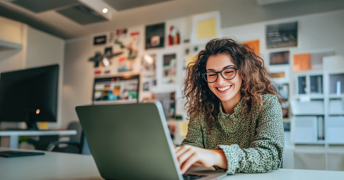A woman with curly hair and glasses smiles while working on her laptop at a desk in a modern, brightly lit office decorated with colorful photos and artwork, as she completes coursework for her online digital marketing degree.