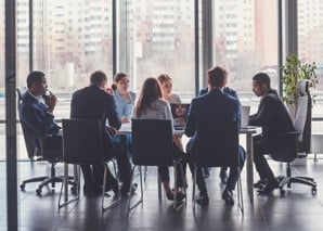 Eight people in business attire sit around a conference table in a modern office with large windows, having a meeting. City buildings are visible outside.