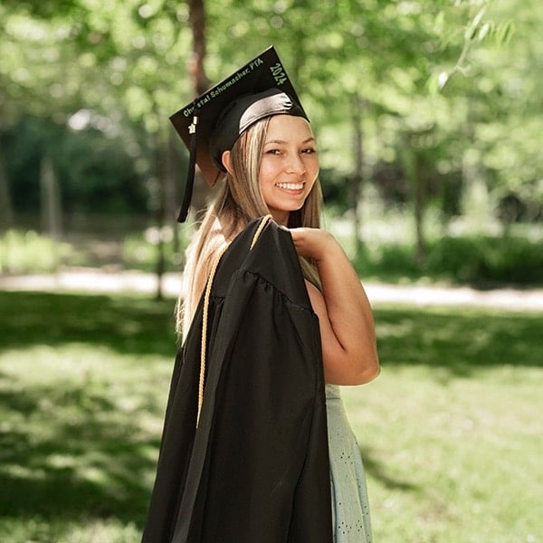 A young woman in a graduation cap and gown smiles over her shoulder while standing outdoors on a sunny day with green trees and grass in the background.