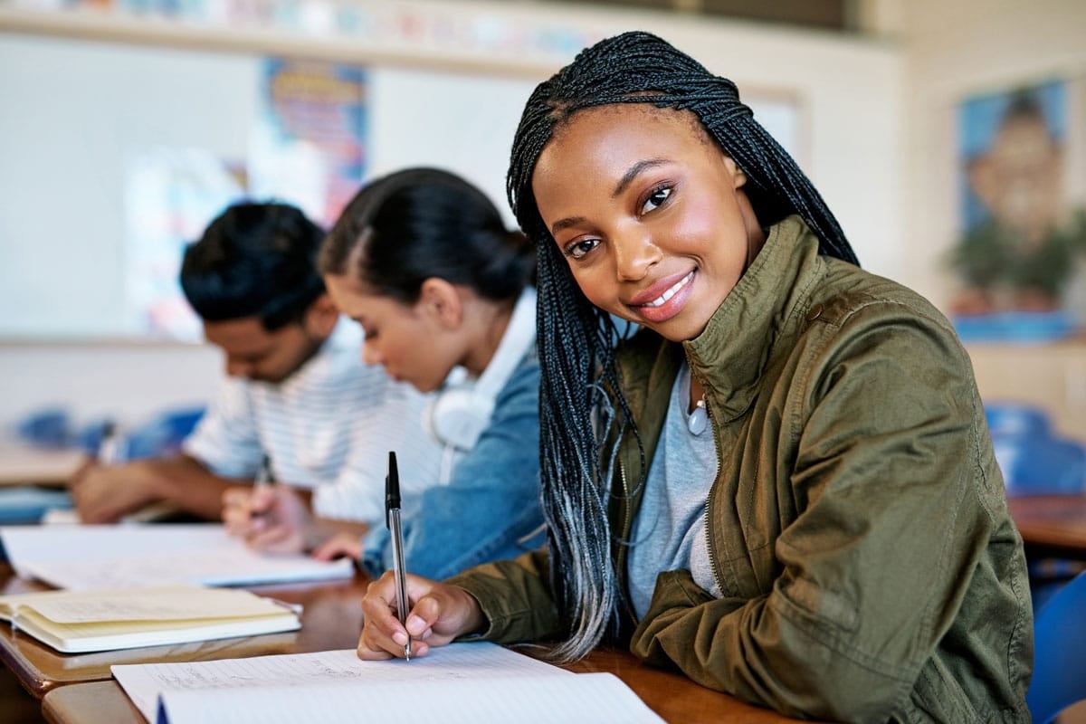 Student with braided hair smiles while taking notes in a classroom; two classmates focus on their work in the background.