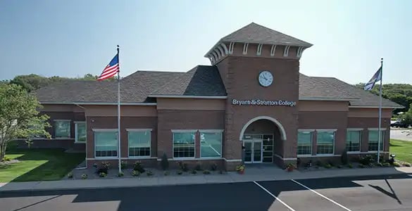 A brick building with "Bryant & Stratton College" signage, two flagpoles, and a clock tower.