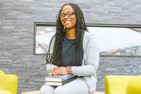 A woman with long braids, wearing glasses and a gray outfit, smiles while sitting indoors. She holds two books on her lap—perhaps researching nursing programs in Virginia Beach—and sits between two green chairs by a gray stone wall.