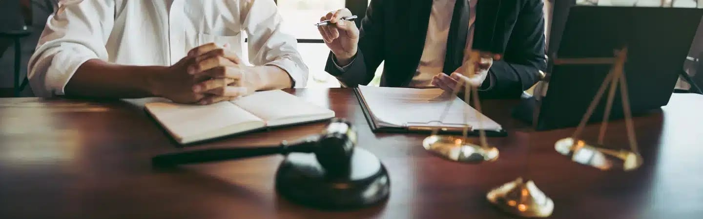 Two individuals sit at a desk with books, legal documents, a gavel, and scales of justice, engaging in a discussion.