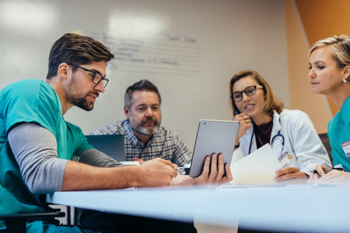Healthcare professionals in a meeting, looking at a tablet and discussing, while seated around a table.