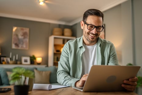 A smiling man wearing glasses sits at a wooden table, using a laptop in a cozy, well-lit living room—perhaps researching business degrees or HR degrees, with shelves and plants in the background.