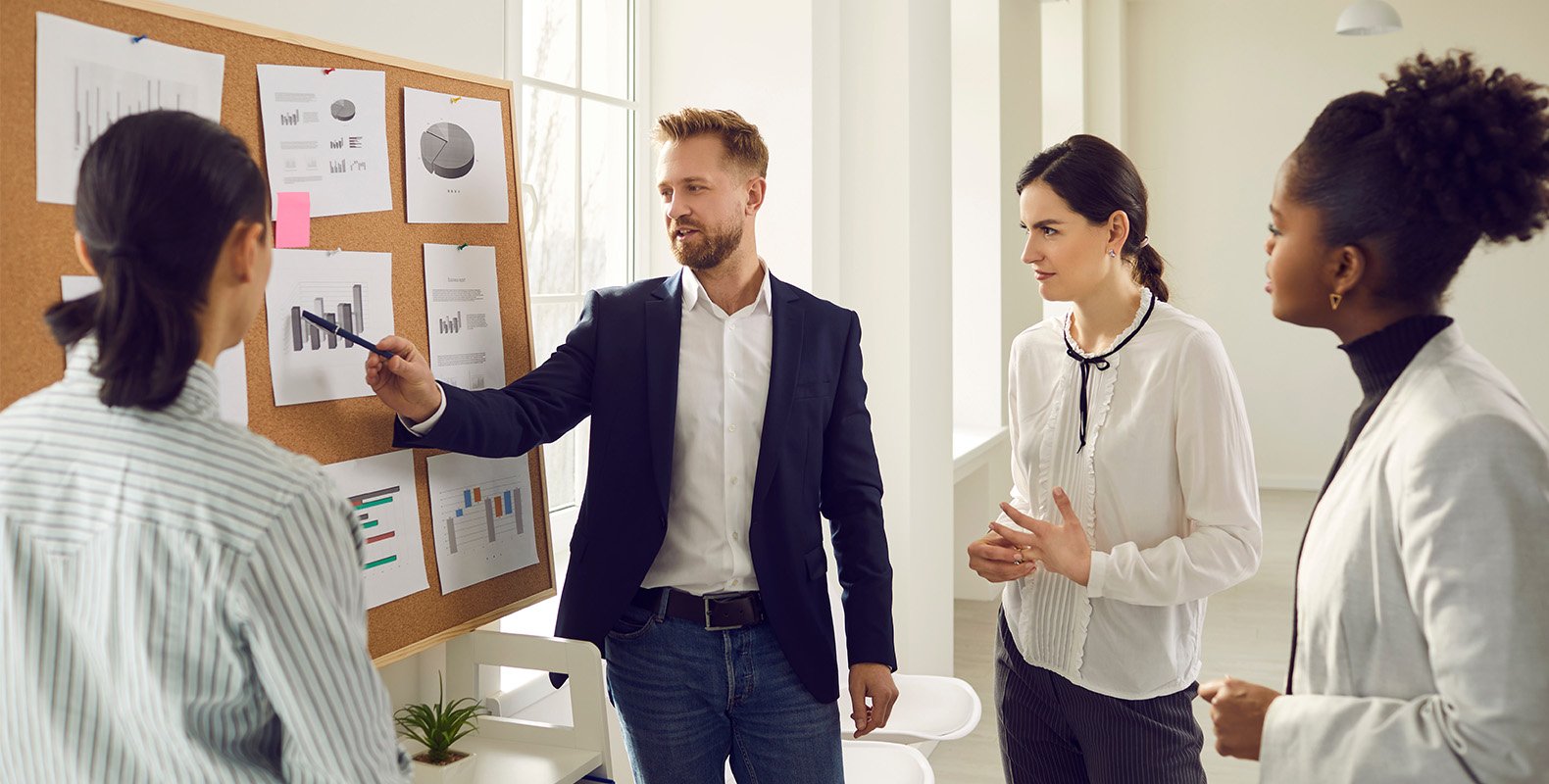 A man in a suit points to charts and graphs on a corkboard while three colleagues, two women and one man, listen attentively in a bright office setting.