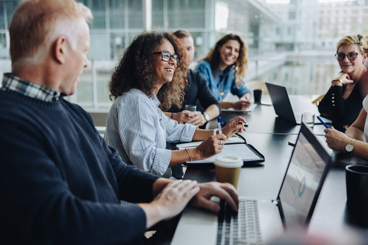 Diverse group of people in a meeting room with laptops and notebooks, discussing and smiling.