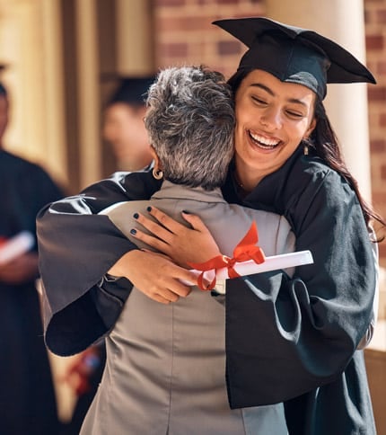 A young graduate in a cap and gown hugs an older person warmly while holding a diploma tied with a red ribbon. They are outdoors, near brick columns, with another graduate visible in the background.