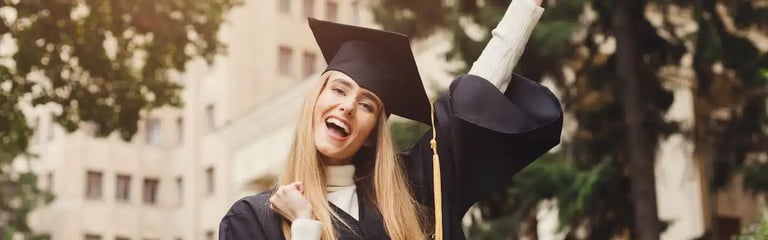 A graduate in a cap and gown celebrates outdoors, holding her diploma and raising her arm joyfully.