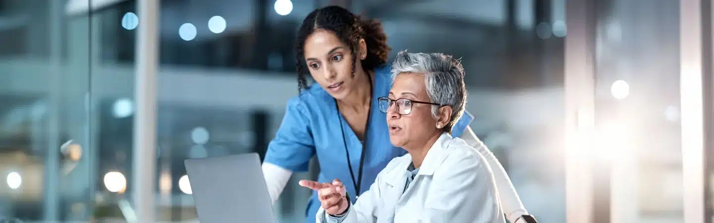 Two healthcare professionals discussing something on a laptop, with one pointing at the screen.
