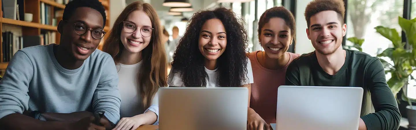 A diverse group of five young adults smiling, sitting with laptops in a bright, modern space.