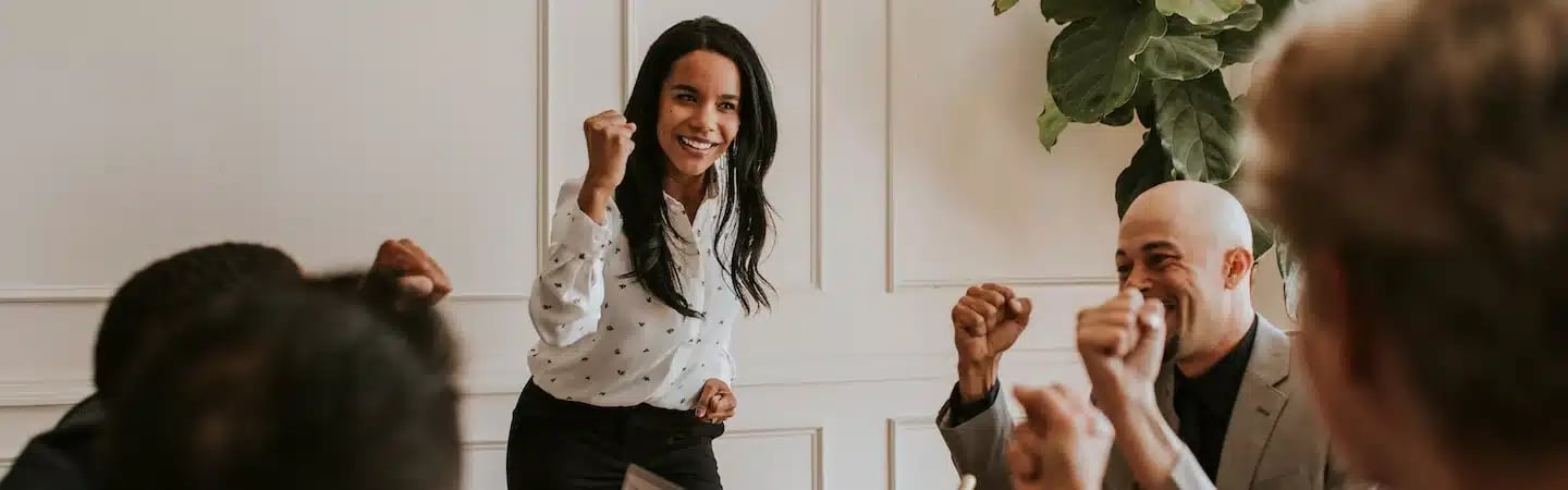 A group of people celebrates in a meeting room. A woman stands in the center with a raised fist, smiling.