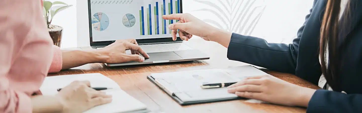 Two people analyzing charts and graphs on a laptop screen and paper documents at a wooden table.