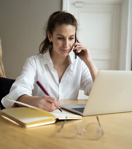 An Administrative Supervisor in a white shirt sits at a table, talking on her phone while writing in a notebook and looking at a laptop. Glasses and another notebook are placed on the table in front of her.