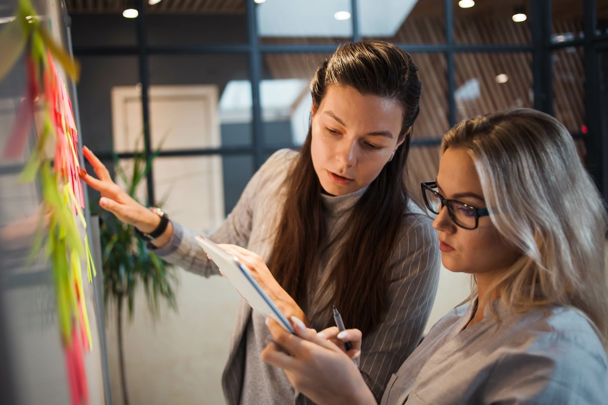 Two women, one holding a tablet, work collaboratively with sticky notes on a glass wall in an office setting.