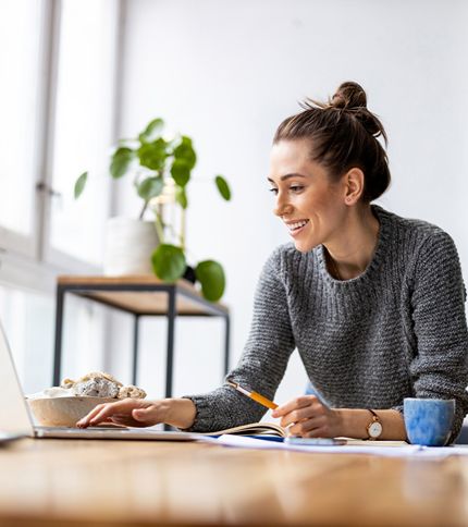 A strategist in a gray sweater sits at a table, smiling while working on her laptop. She holds a pencil with her notebook and coffee cup nearby, ready to tackle SEO tasks. A plant is visible on a shelf in the background.