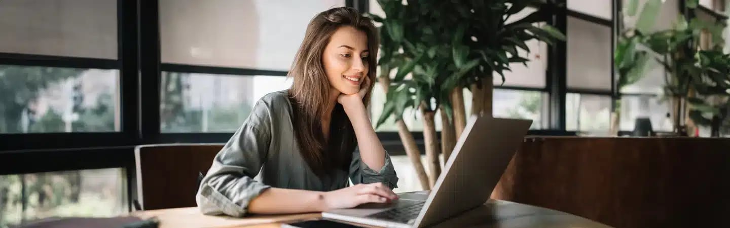 A woman is sitting at a table, smiling and using a laptop in a bright room with large windows and plants.