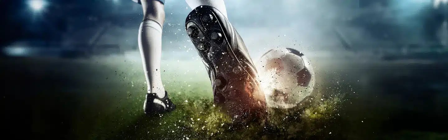 Close-up of a soccer player's leg and foot, wearing cleats, kicking a soccer ball on a wet, grassy field at night.