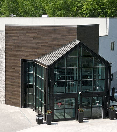 Entrance of Bryant & Stratton College Campus in Liverpool, NY with large glass panels forming a triangular facade, dark metal framing, and wood-textured wall accents. Several small flower pots are placed near the glass doors in this NY-inspired design. Trees are visible in the background.