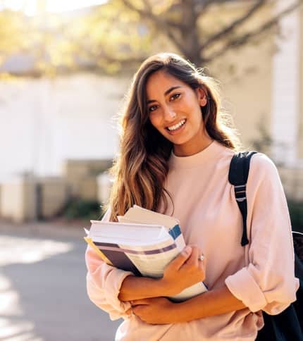 A young woman is outdoors, smiling, and holding several books. She wears a light pink sweater and a backpack. The background is softly focused, suggesting a sunny day on a campus or park setting.