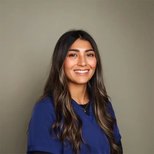 A woman with long, wavy brown hair smiles at the camera. She is wearing navy blue scrubs and standing in front of a plain, light olive background.