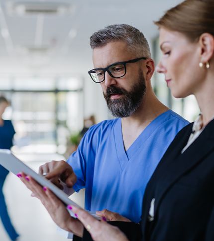 A male healthcare professional in blue scrubs and glasses discusses information on a tablet with a Medical Office Manager in business attire in a bright hospital corridor.