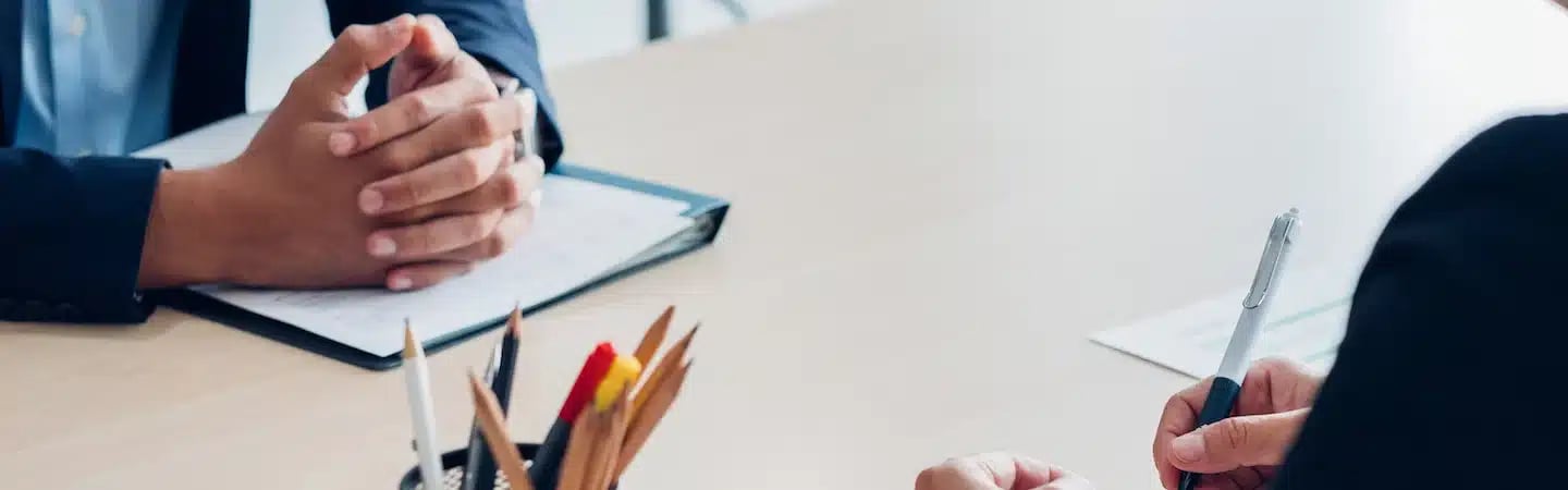 Two people seated at a table during a meeting, with one person holding a pen and the other with hands clasped.