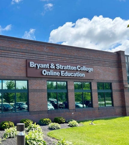 Modern brick building exterior with a sign reading 'Bryant & Stratton College' and a clear blue sky with clouds.