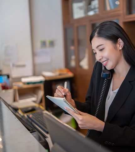 A woman in a black blazer, likely a Hotel Desk Clerk, is talking on the phone and smiling while writing on a notepad at an office or reception desk, working professionally with computers and shelves behind her.