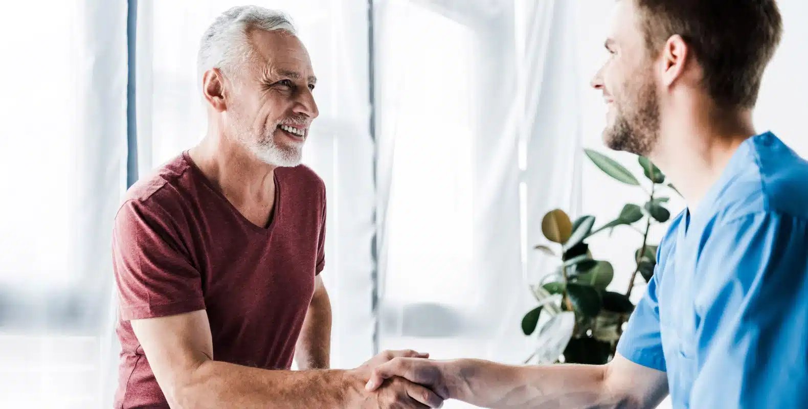 An older man shakes hands with a healthcare professional in a bright room. Both are smiling.