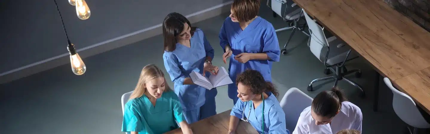 Group of medical professionals in scrubs gathered around a table, discussing and consulting documents.