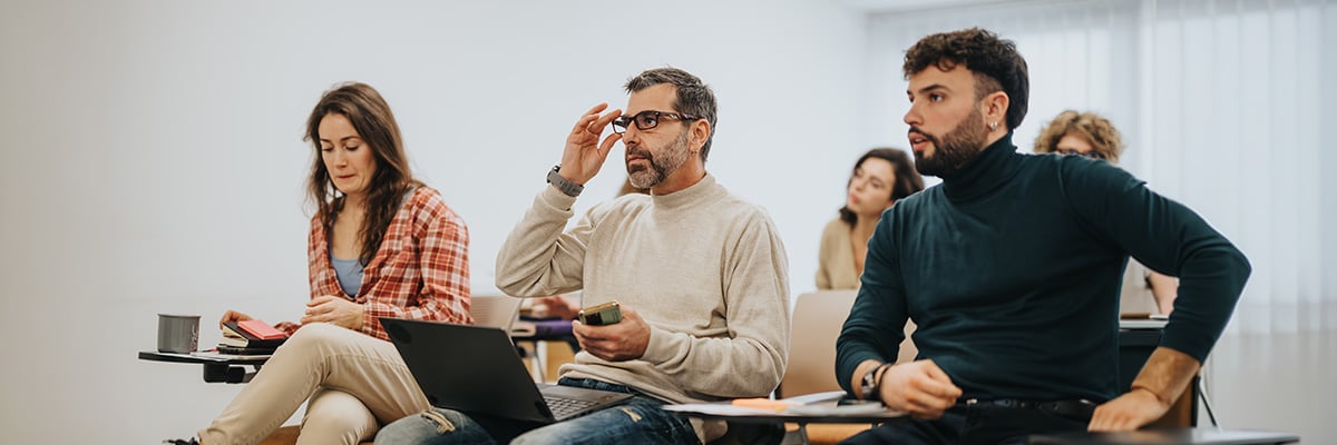 A group of adults sit in a classroom setting, engaged in continuing education. One man adjusts his glasses, while others with notebooks and laptops listen attentively. The atmosphere is focused and motivated to learn.