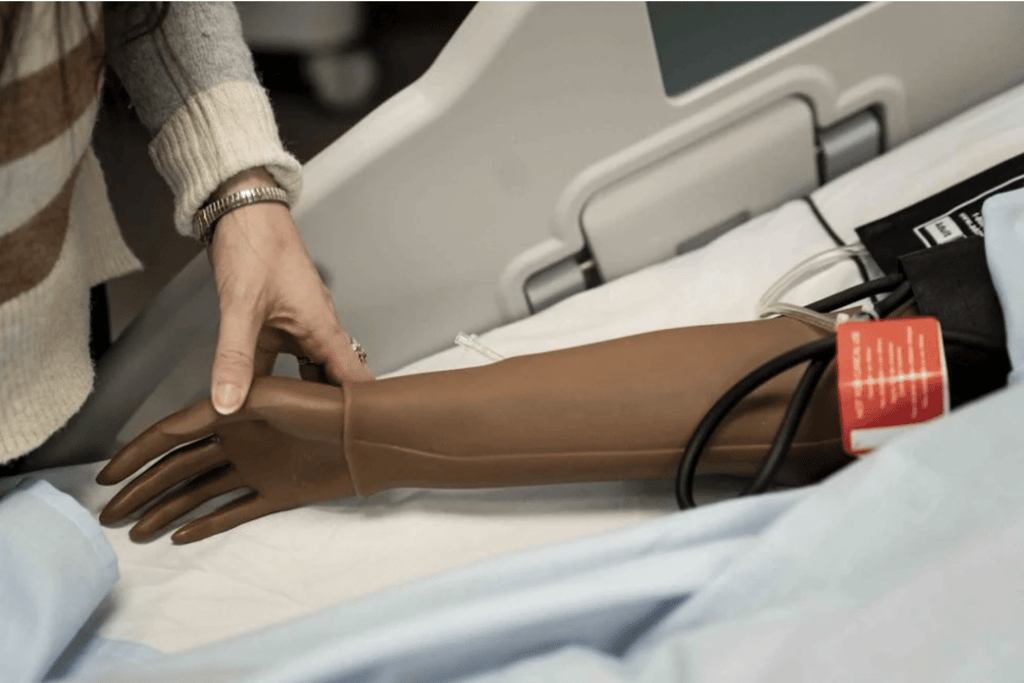 A person adjusts a brown prosthetic arm lying on a hospital bed, next to medical equipment and partially covered by a blue sheet.