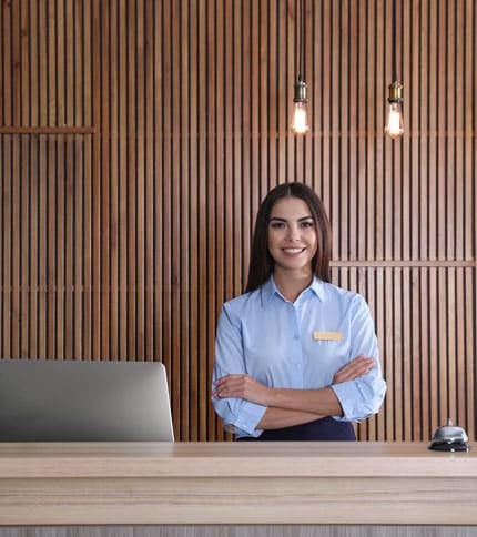 A smiling woman in a blue uniform stands with arms crossed behind a reception desk. A laptop and bell are on the desk. Wood panel wall and hanging light bulbs are in the background.