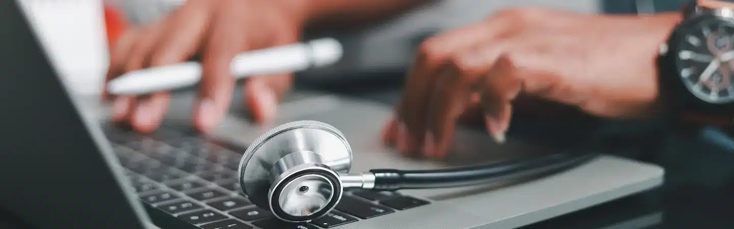 A person types on a laptop with a stethoscope lying on the keyboard, suggesting medical work or telemedicine.