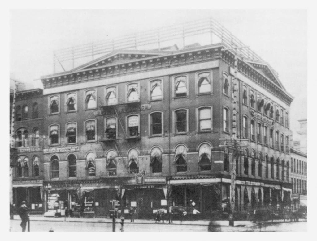 Historic black-and-white photo of a four-story brick building with ground-floor shops and businesses in an urban area.