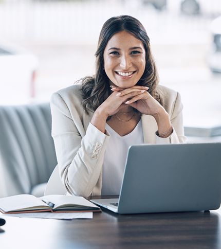 A Legal Office Assistant in a light blazer smiles while sitting at a desk with a laptop and an open notebook, resting her chin on her hands in a bright, modern office setting.