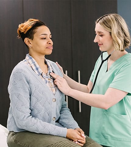 A healthcare professional in green scrubs uses a stethoscope to check the heartbeat of a patient sitting on an examination table. The patient is wearing a light blue cardigan and a checkered shirt. They are in a medical office setting.