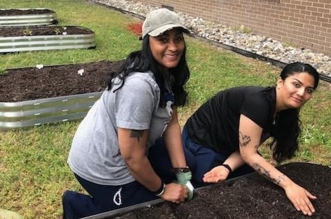 Two women smile while tending to raised garden beds outside on a grassy area. They are planting seeds in the soil.