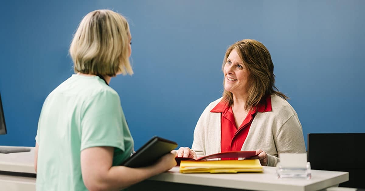 Woman in red shirt and white cardigan speaking with a nurse at a reception desk. They both have open folders.