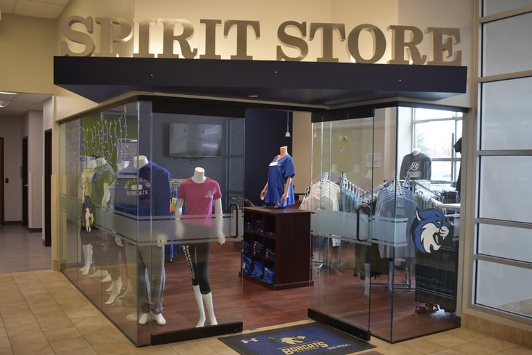 Entrance to a "Spirit Store" with sports apparel on display, mannequins dressed in team jerseys, and a plush mascot mat.