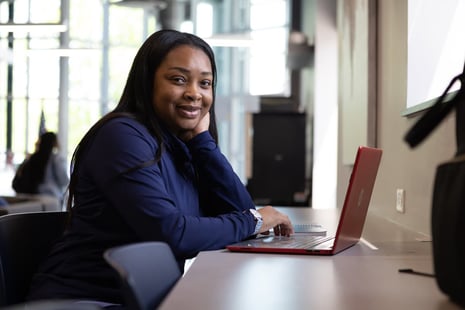 A woman sits at a desk with a red laptop, smiling at the camera. She rests her head on one hand and appears to be working in a bright, modern indoor space.