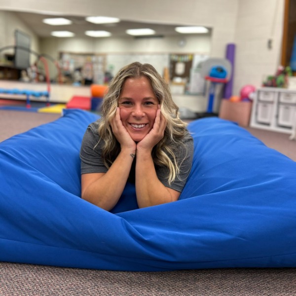 A woman with blonde hair smiles while lying on her stomach on a large blue beanbag in a brightly lit room, possibly a classroom or play area.